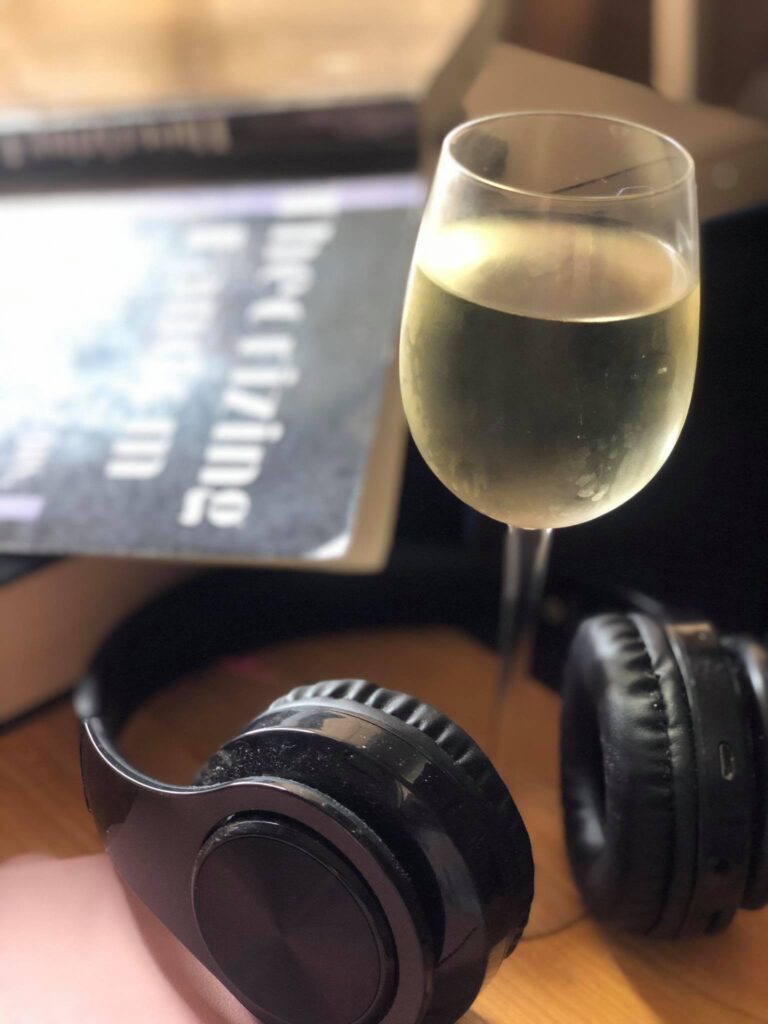 Wine glass with white wine on a desk with black headphones and an open book. Book is face down and titled Theorizing Fandoms